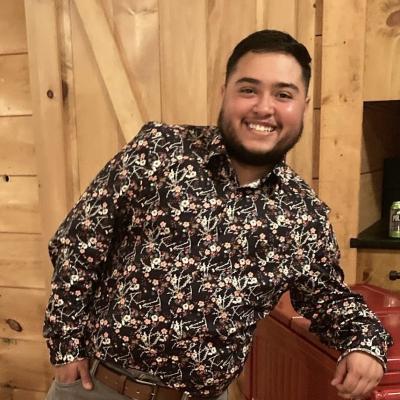 Maureo is pictured, smiling, in a brown patterned shirt against a wood backdrop.