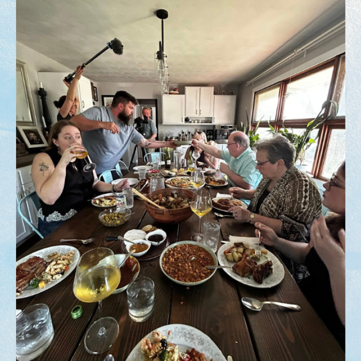 A dozen people sit for dinner at a home, with a small film crew setting up equipment around the room.